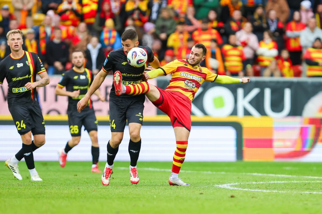 Kadr z meczu Jagiellonia Białystok - Korona Kielce Dwóch piłkarzy rywalizuje o piłkę na boisku podczas meczu, jeden z nich unosi nogę do wysokości rywala, w tle widownia ubrana w klubowe barwy.