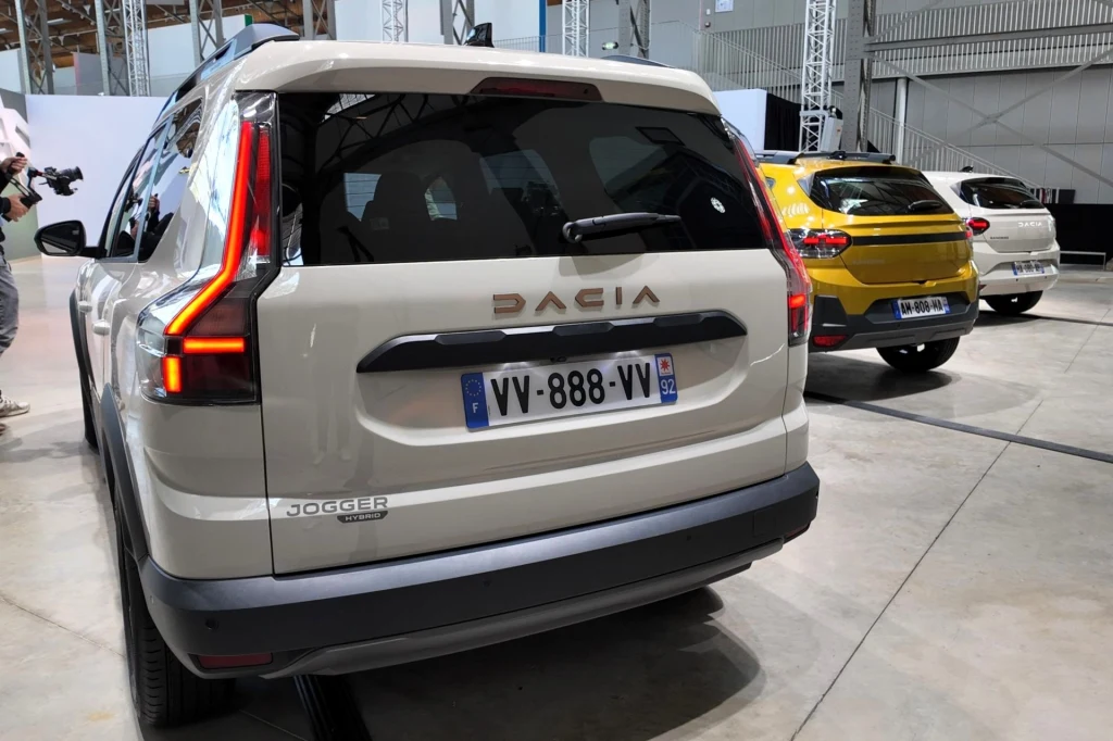 Nowe tylne światła Dacii Jogger również nawiązują do litery "T". Tylna część beżowego samochodu marki Dacia Jogger z widocznym logo i tablicą rejestracyjną, z przodu po lewej stronie znajduje się żółty samochód tej samej marki, całość ustawiona w hali wystawowej.