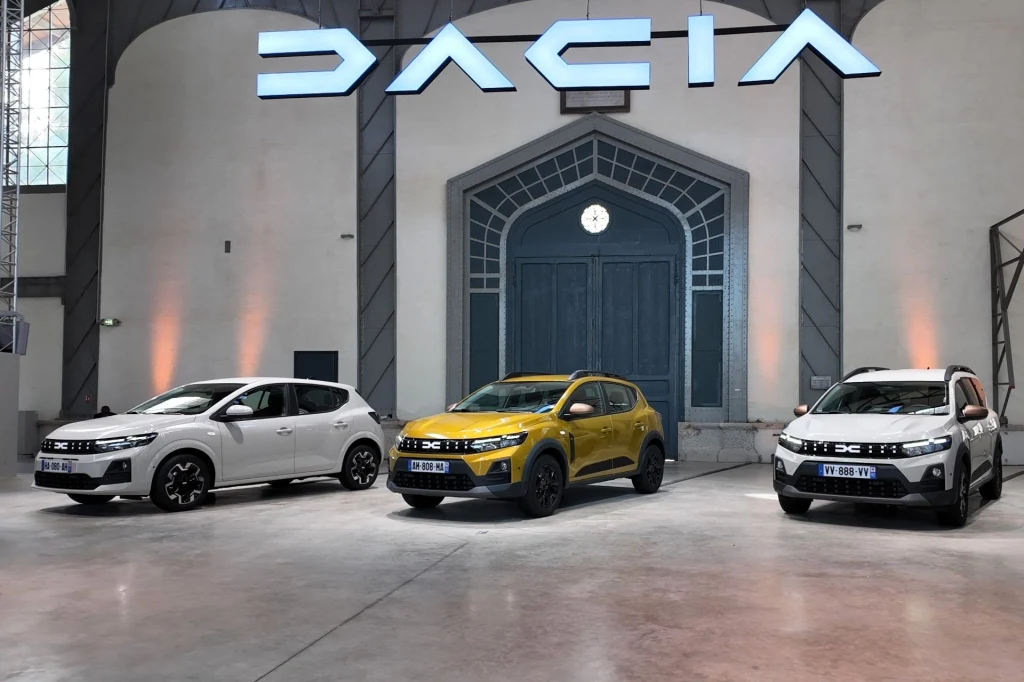 Dacia Sandero i Jogger to trzon sprzedaży rumuńskiej marki. Pierwszy z modeli zdobył tytuł najchętniej kupowanego samochodu w 2024 roku w Europie. Trzy nowe samochody marki Dacia, ustawione w przestronnej, dobrze oświetlonej hali wystawienniczej, na pierwszym planie żółty crossover, po bokach dwa białe modele, nad nimi duży napis DACIA.