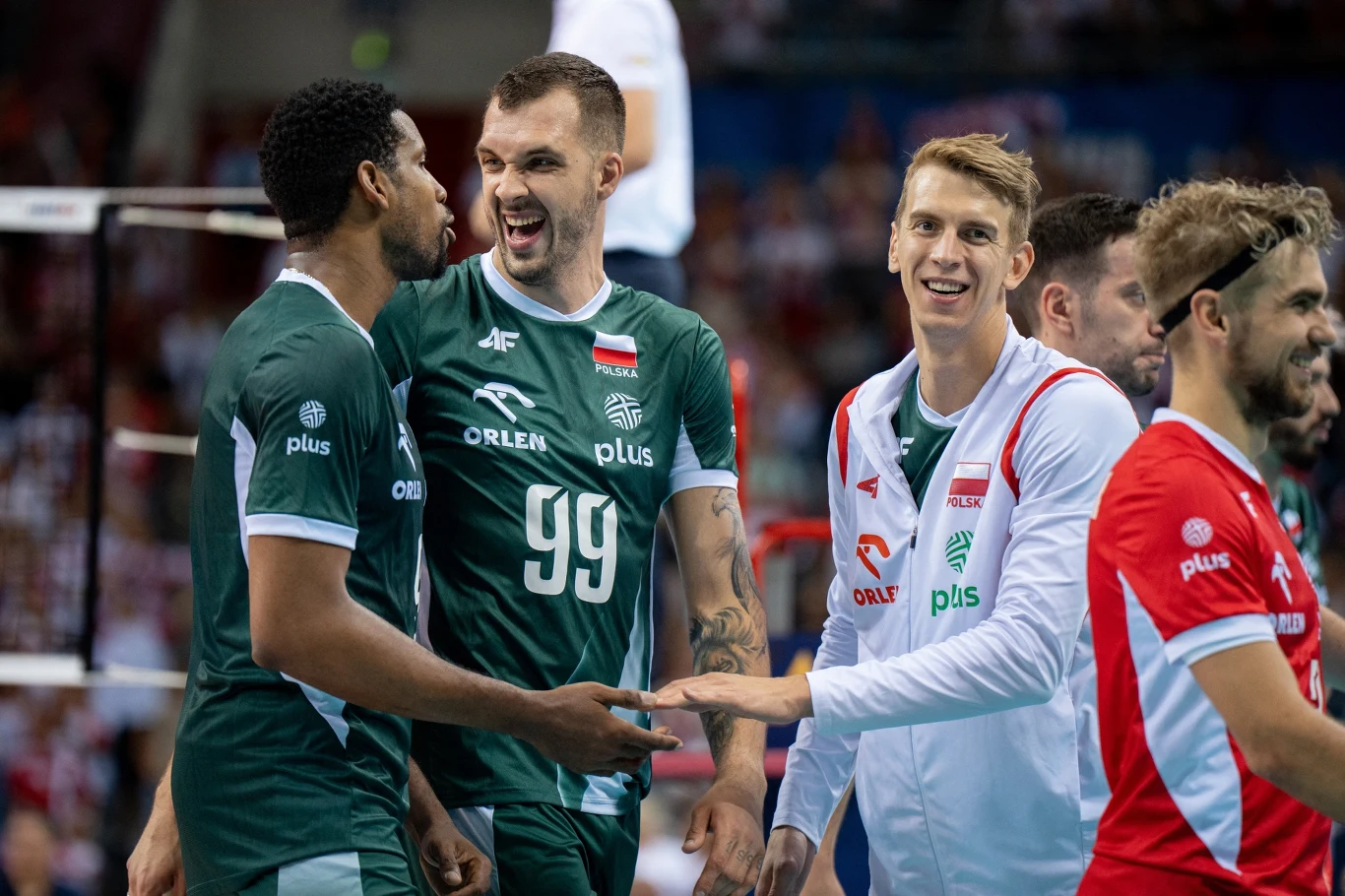 Norbert Huber (z numerem 99) w reprezentacji Polski Grupa siatkarzy w zielonych i białych strojach celebruje punkt lub zwycięstwo, uśmiechnięci zawodnicy gestykulują i patrzą na siebie, w tle widoczni inni członkowie drużyny oraz publiczność.