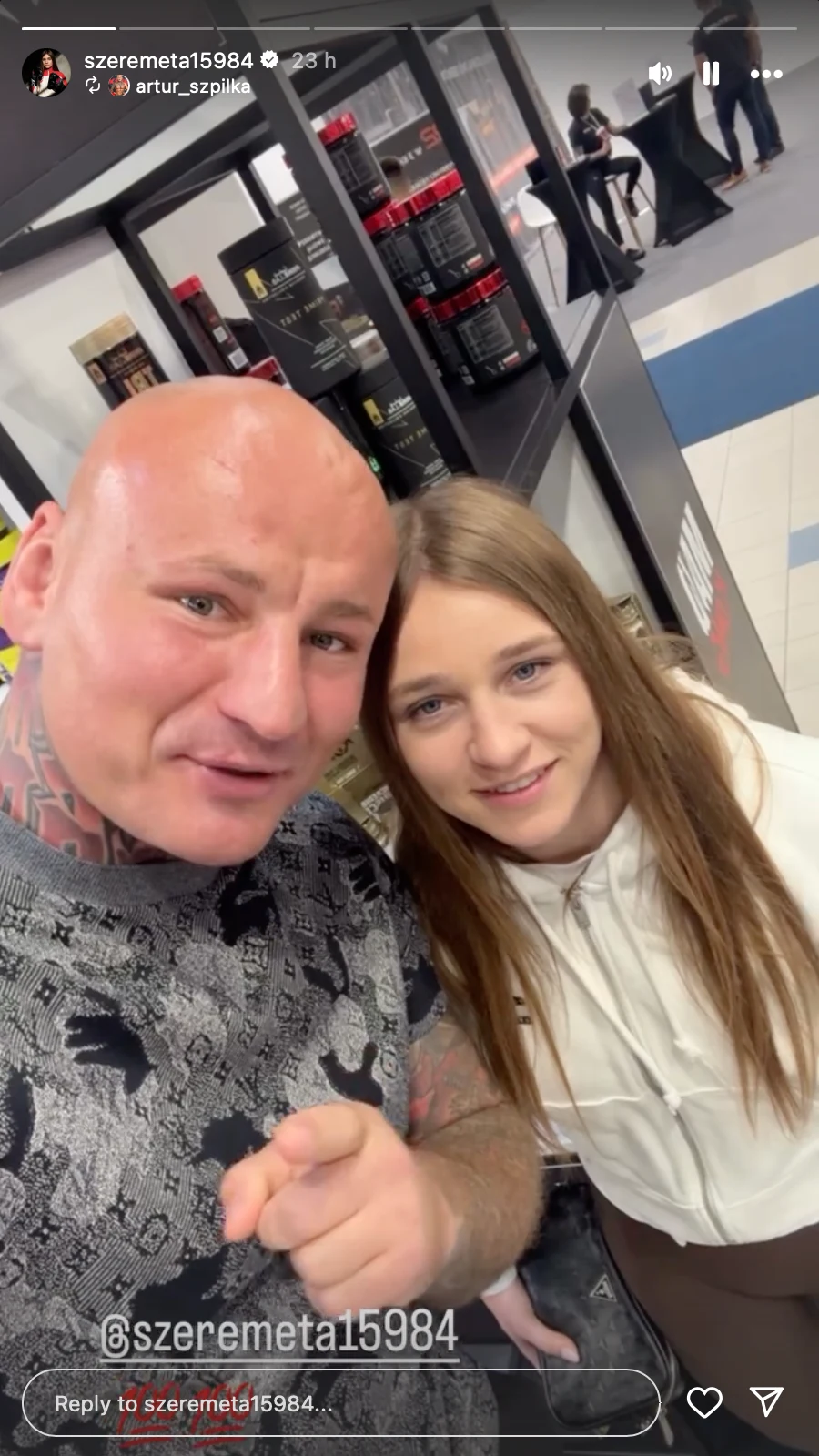 Mężczyzna z widocznymi tatuażami na karku i głowie oraz młoda kobieta stoją razem w sklepie z suplementami i produktami sportowymi, patrząc i uśmiechając się do aparatu.