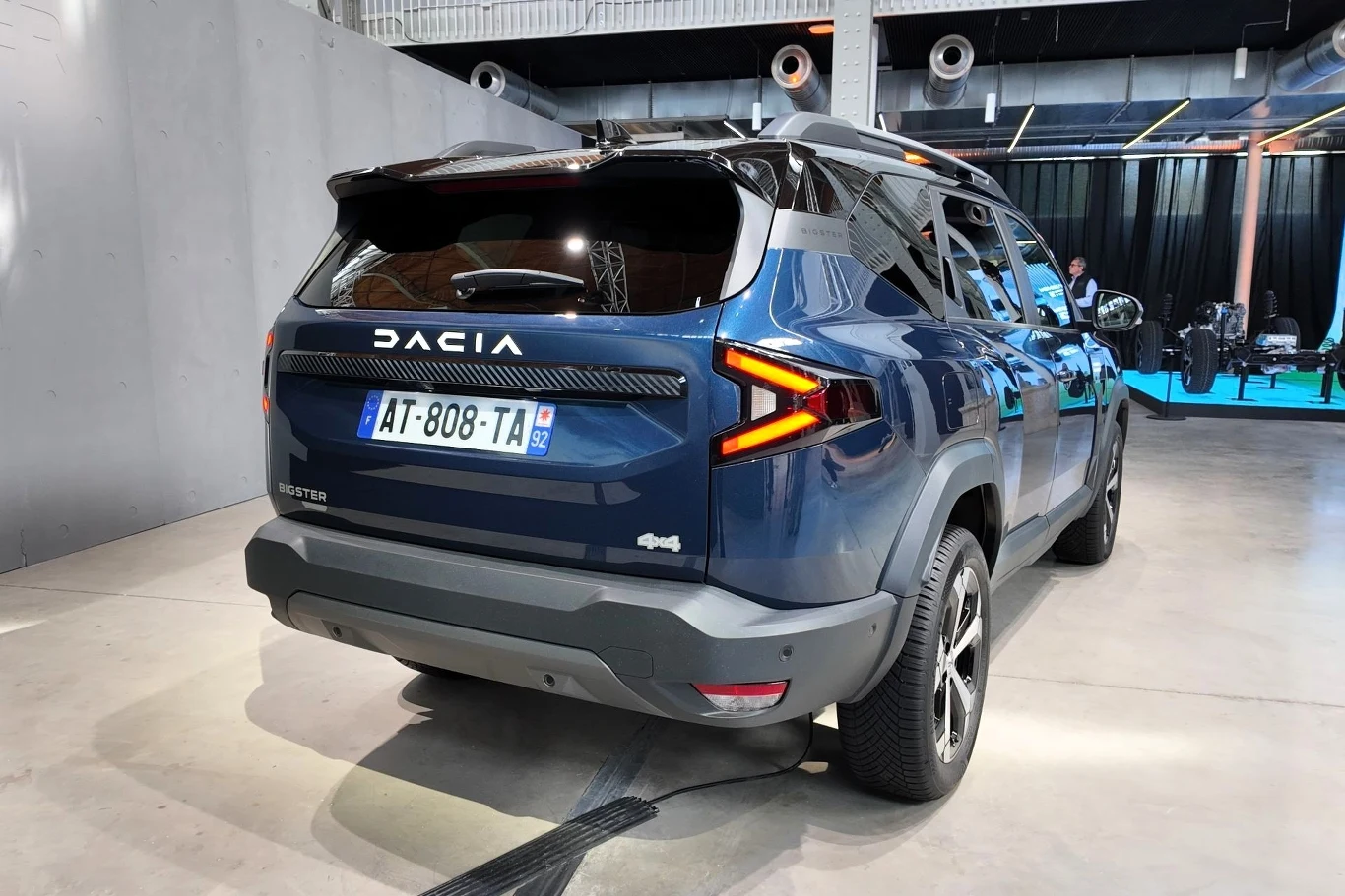 Granatowy SUV marki Dacia z francuskimi tablicami rejestracyjnymi stoi w przestronnej hali na betonowej podłodze; samochód zwraca uwagę nowoczesnymi, wyrazistymi tylnymi lampami i masywną sylwetką.