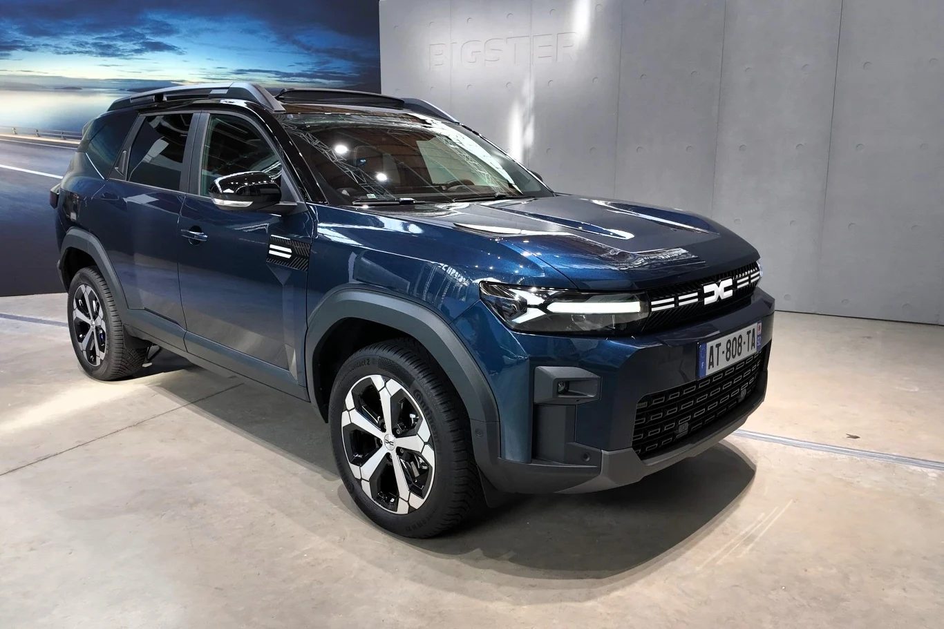 Nowoczesny samochód osobowy typu SUV o sportowym wyglądzie, niebieski lakier metalik, charakterystyczny grill i wyraziste reflektory, pojazd stoi w jasnym, minimalistycznym wnętrzu, na tle ściany i częściowo widocznej grafiki z motywem drogi.