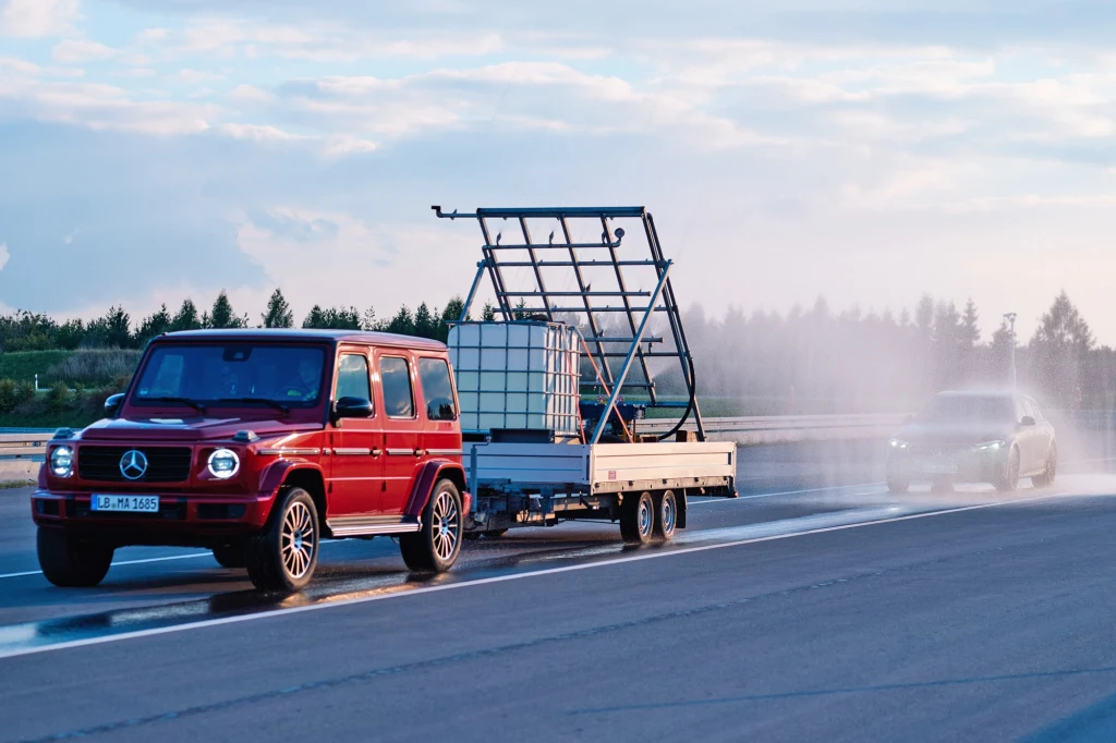 Tak wygląda instalacja do tworzenia sztucznego deszczu, umieszczone na przyczepie ciągniętej przez klasę G Czerwony Mercedes G z przyczepą transportującą zbiornik plastikowy oraz metalową ramę, poruszający się po mokrej nawierzchni, w tle inne auto i rozpryskująca się woda.