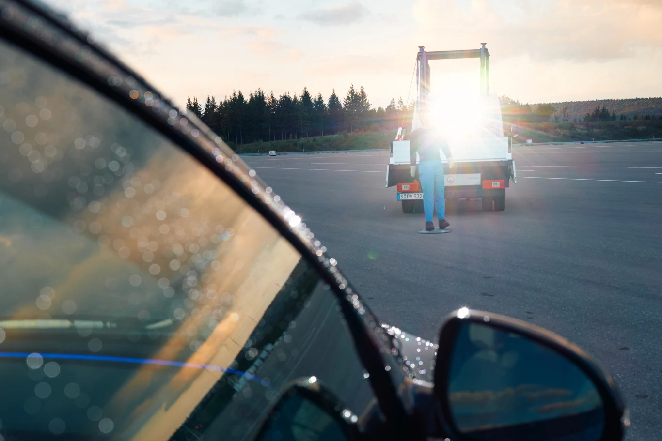 Auto na pierwszym planie z widocznymi kroplami deszczu na szybie oraz laweta z osobą stojącą obok na pustym placu otoczonym lasem, światło odbijające się od lawety tworzy silny efekt świetlny.
