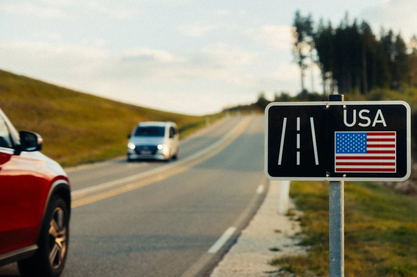 Droga prowadząca przez tereny zielone, po której poruszają się samochody, przy drodze stoi znak z flagą USA oraz napisem 'USA', w tle widoczny las.