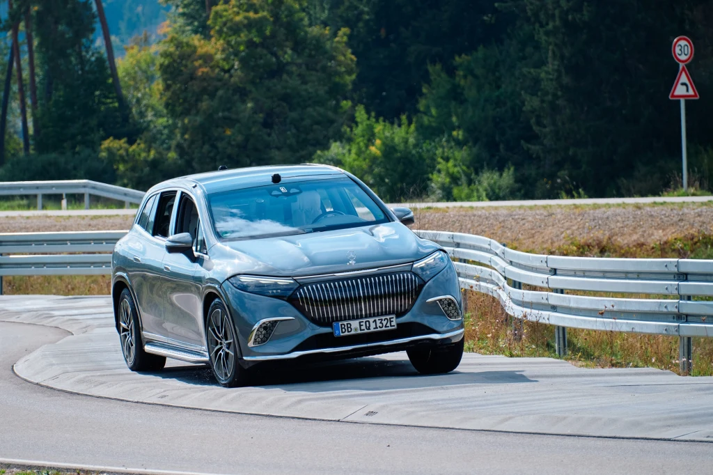 Zdjęcie tego nie oddaje, ale samochody przejeżdżające przez ten fragment dosłownie podskakują na wybojach. Gdyby w środku ktoś siedział, byłoby to bardzo nieprzyjemne doświadczenie Nowoczesny srebrny samochód osobowy na zakręcie drogi testowej, otoczony metalowymi barierkami ochronnymi, w tle zielone drzewa oraz znak ostrzegawczy o niebezpiecznym zakręcie.