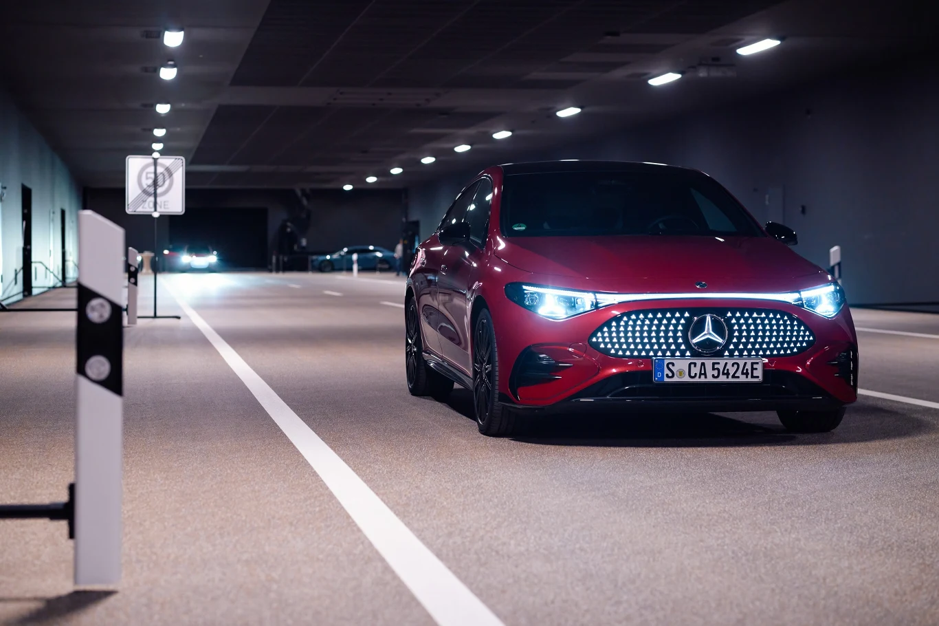 czerwony samochód marki Mercedes-Benz jadący oświetlonym tunelem, wyraźnie widoczne światła przednie oraz tablica rejestracyjna pojazdu, w tle inne pojazdy i elementy infrastruktury drogowej jak linie oraz znaki