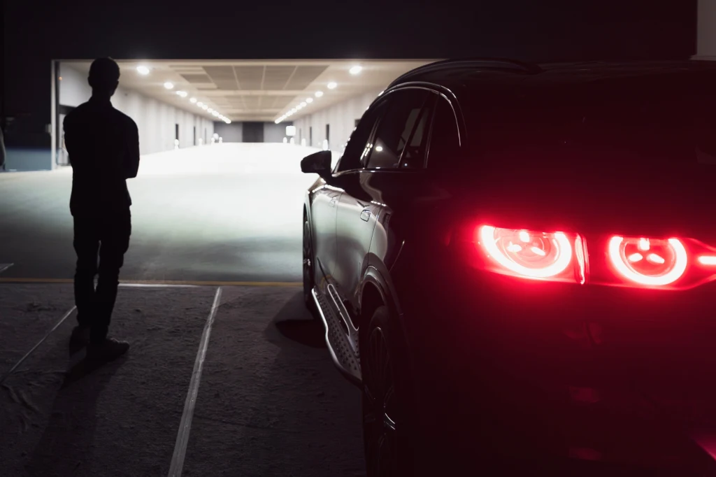 Odcinek drogi do testowania oświetlenia ma 135 m długości Sylwetka osoby stojącej obok czarnego samochodu z włączonymi tylnymi światłami, naprzeciwko jasnego wejścia do dużego budynku na tle nocnej scenerii.