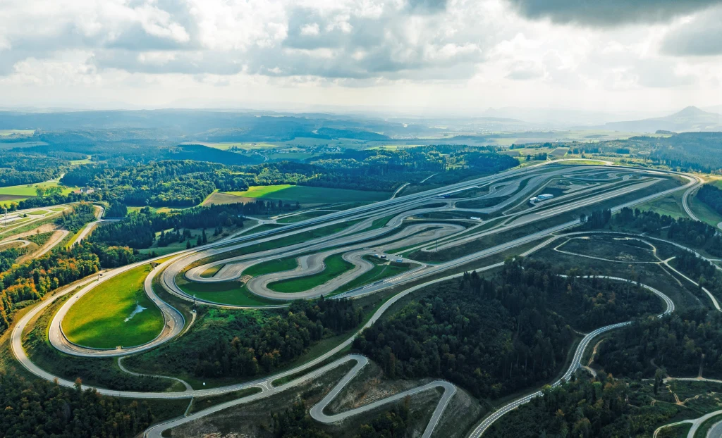 Immendingen Test and Technology Centre robi wrażenie swoimi rozmiarami i liczbą dostępnych dróg Kręty tor wyścigowy otoczony zielenią, położony wśród pagórków i lasów, z widoczną z lotu ptaka całą infrastrukturą oraz malowniczym krajobrazem w tle.