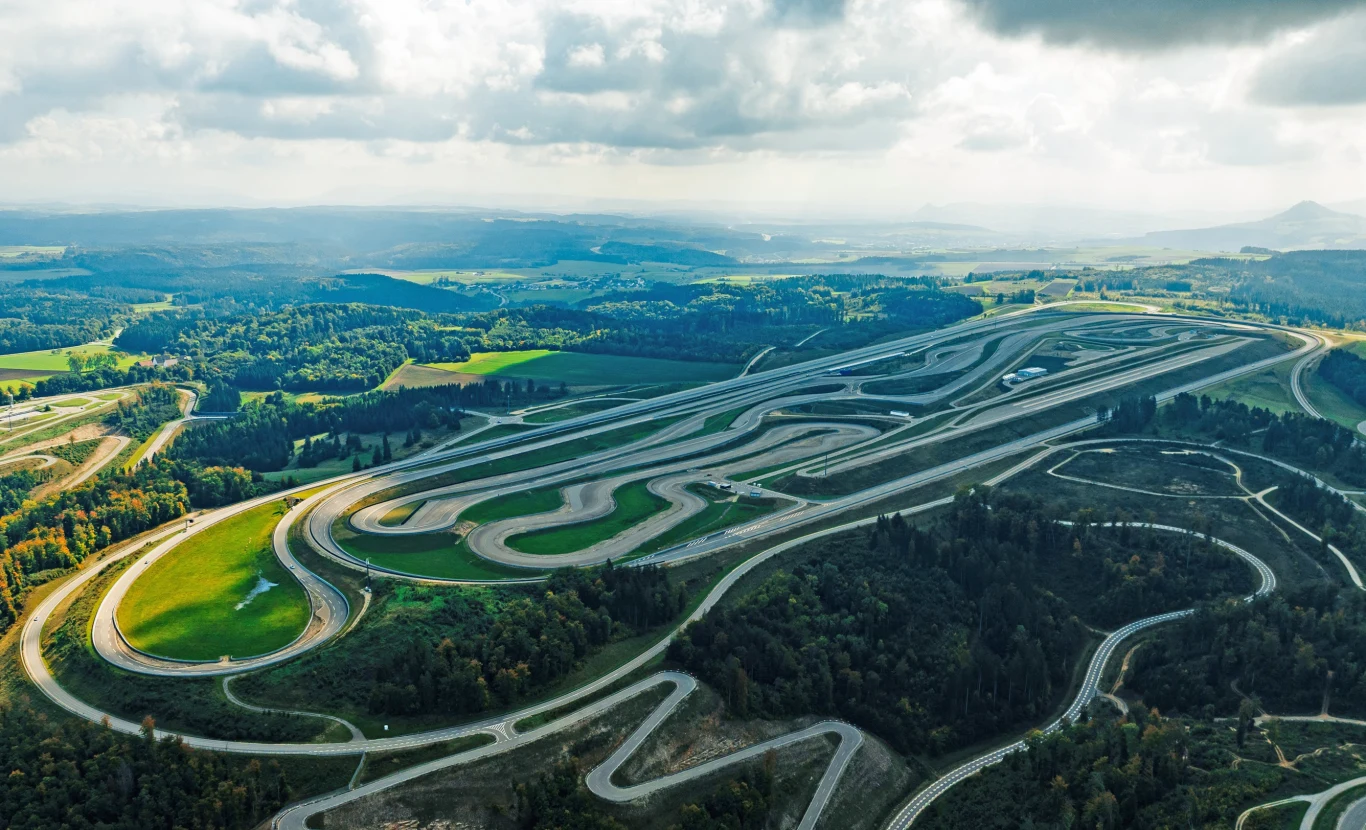 Kręty tor wyścigowy otoczony zielenią, położony wśród pagórków i lasów, z widoczną z lotu ptaka całą infrastrukturą oraz malowniczym krajobrazem w tle.