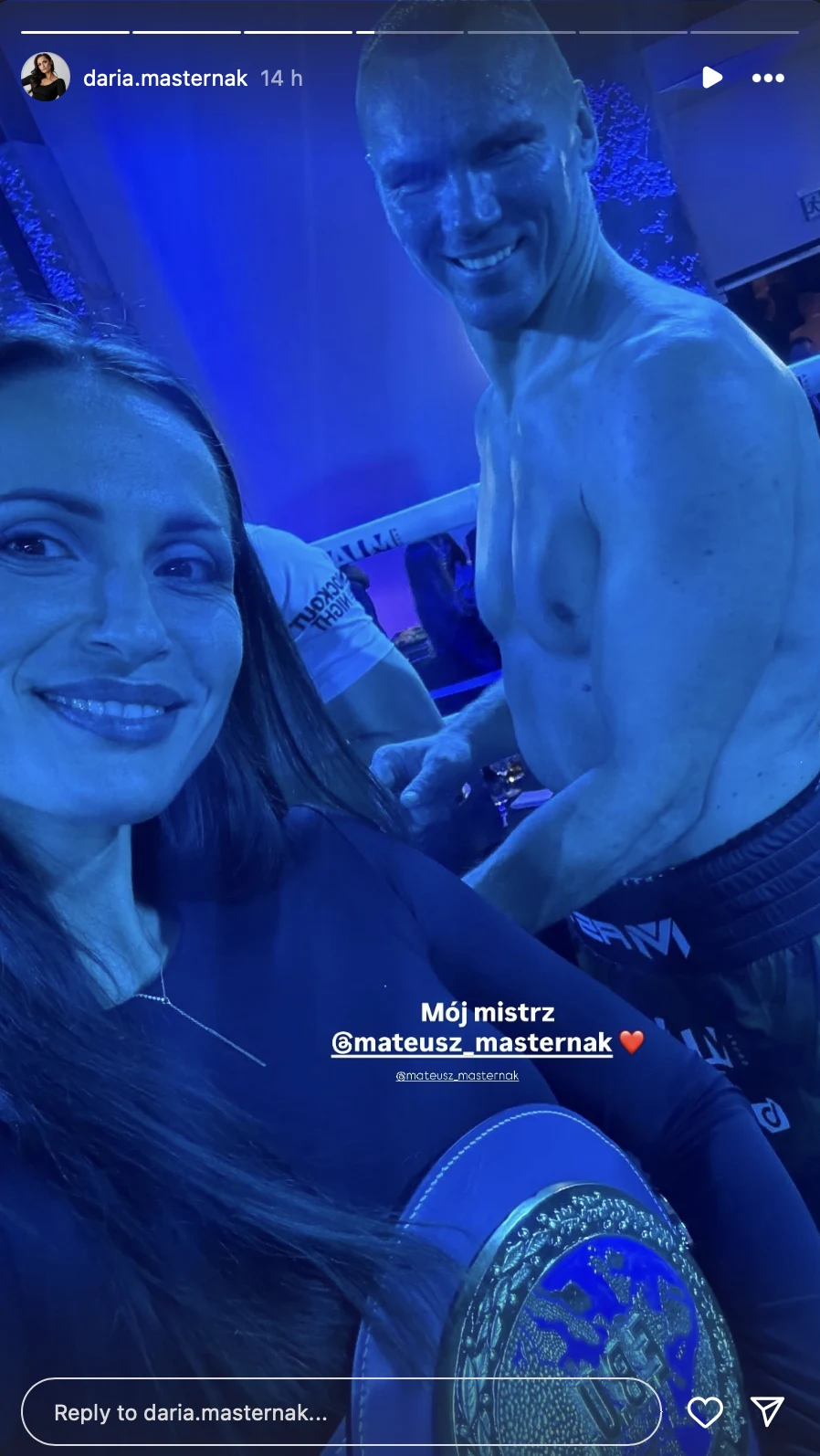 Daria Masternak zabrała głos po triumfie męża w Zakopanem Kobieta uśmiecha się do aparatu, trzymając pas mistrzowski, obok znajduje się uśmiechnięty bokser bez koszulki, na który również skierowana jest kamera. Oświetlenie ma niebieski odcień, a atmosfera wskazuje na radosny moment po walce bokserskiej.