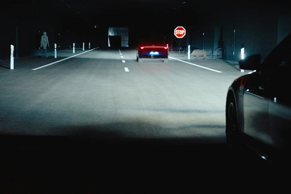 Inne pojazdy, znaki, piesi, dzikie zwierzęta - w centrum testowania oświetlenia Mercedesa można aranżować skomplikowane sytuacje drogowe Jazda samochodami w ciemnym tunelu, widoczne tylne światła pojazdu oraz znak stop i sylwetka ludzkiej figury po lewej stronie drogi.