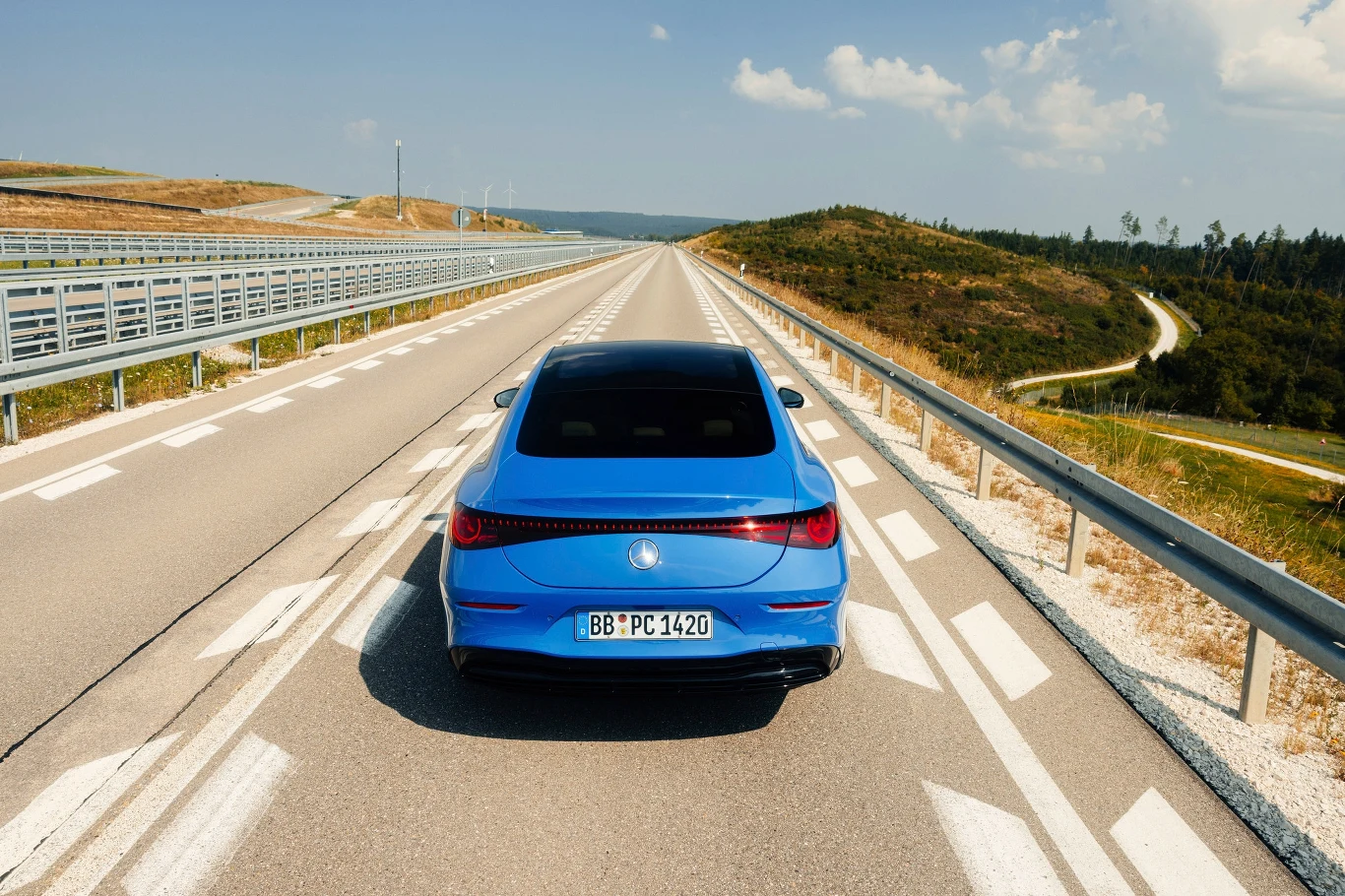 Niebieski samochód jedzie pustą autostradą otoczoną przez zielone wzgórza i niebo z lekkim zachmurzeniem