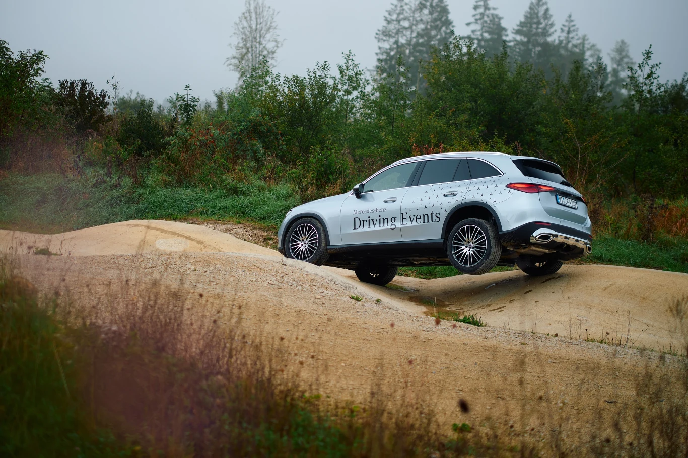 Nowoczesny srebrny SUV z napisem Driving Events pokonuje wyboisty, piaszczysty teren, otoczony zielenią i drzewami w tle, koła uniesione nad ziemią prezentują możliwości terenowe pojazdu.