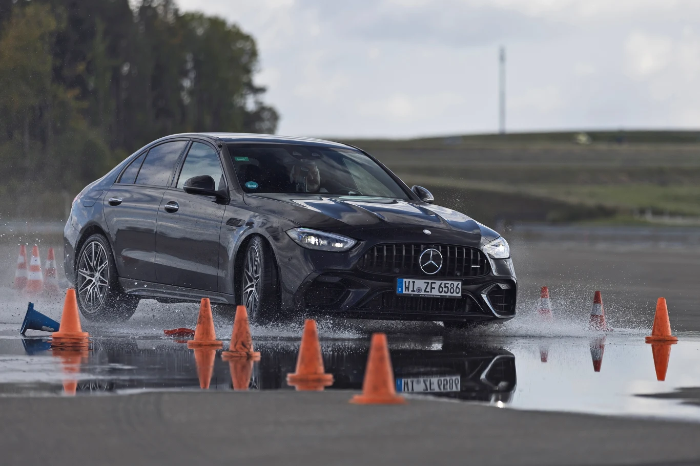 Czarny samochód marki Mercedes wykonuje dynamiczny manewr na mokrej nawierzchni, otoczony pomarańczowymi pachołkami podczas testu na torze.