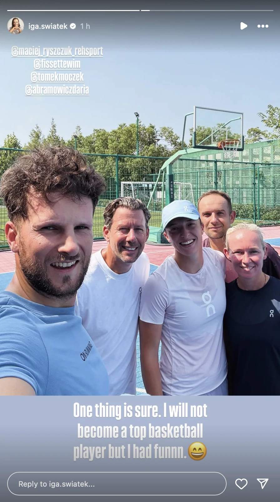 Iga Świątek i jej team na koszykarskim parkiecie Grupa pięciu uśmiechniętych osób pozujących wspólnie na boisku do koszykówki w słoneczny dzień, w tle widoczny kosz oraz zielone ogrodzenie.