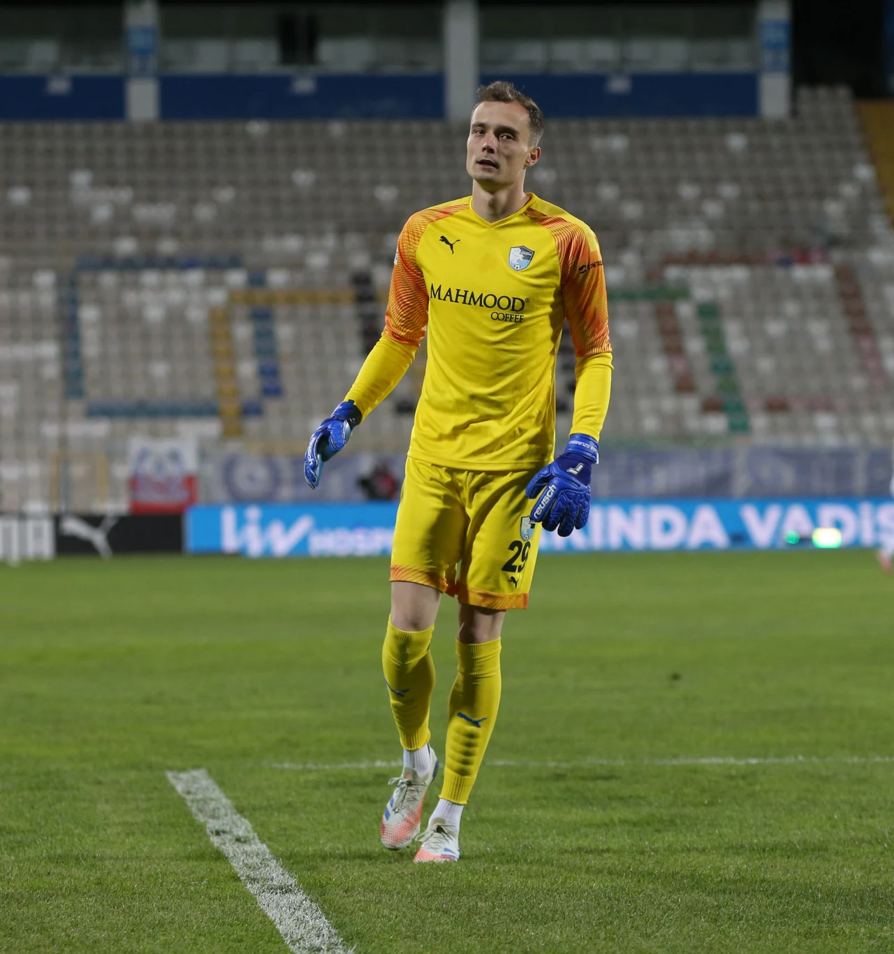 Jakub Szumski Bramkarz w żółtym stroju z niebieskimi rękawicami stoi na murawie stadionu piłkarskiego, w tle widoczne są puste trybuny i fragmenty reklam.