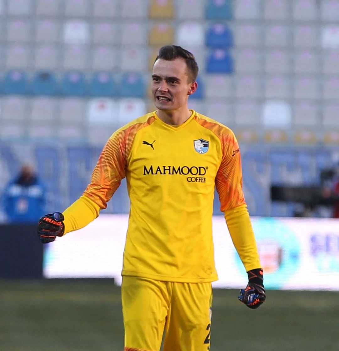 Jakub Szumski Piłkarz w stroju bramkarza w żółtym komplecie z rękawicami stoi na tle pustych trybun stadionu, na jego koszulce znajduje się logo sponsora oraz herb drużyny.