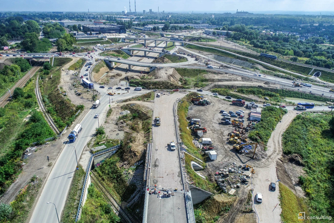 Poziomo biegnie ulica Kocmyrzowska, a pionowo Łowińskiego oraz budowany odcinek S7. W tle widać nowohucki Kombinat Plac budowy dużego węzła drogowego z wieloma rozwidleniami i maszynami budowlanymi, pojazdami oraz rozkopanym terenem, otoczony zielenią i drogami dojazdowymi, widoczne prace ziemne i inżynieryjne.
