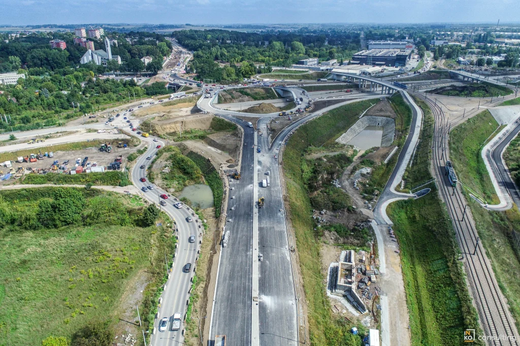 Widoczna pośrodku dwupasmowa droga to docelowy ślad ulicy Kocmyrzowskiej Rozległa budowa nowoczesnego węzła drogowego z licznymi pasami ruchu, pojazdami budowlanymi oraz otaczającą infrastrukturą kolejową i zielenią, w tle widoczne budynki mieszkalne i przemysłowe.