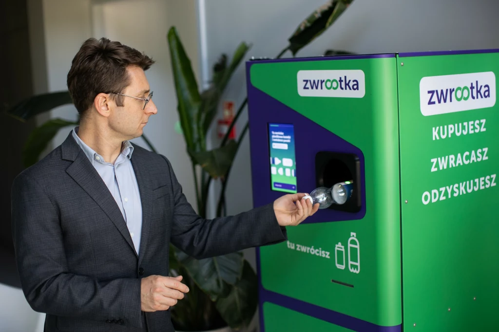 Mężczyzna w okularach i garniturze wkłada plastikową butelkę do automatu zwrotnego oznaczonego napisem 'zwrótka' w nowoczesnym wnętrzu biurowym, w tle zielone rośliny.