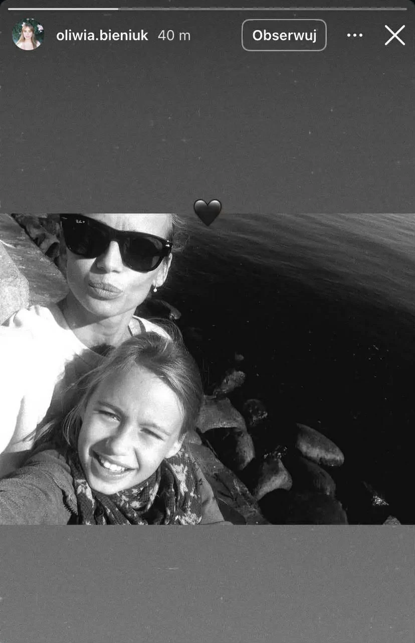 Dwie uśmiechnięte osoby, jedna w okularach przeciwsłonecznych, robią wspólne selfie na tle kamieni i ciemnej tafli wody, w tle widoczne skały.