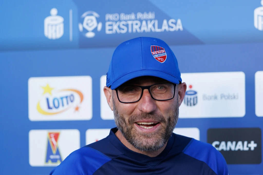 Mężczyzna w sportowym stroju i czapce z daszkiem, z brodą oraz okularami, uśmiecha się, stojąc na tle tablicy z logotypami sponsorów oraz napisami Ekstraklasa.