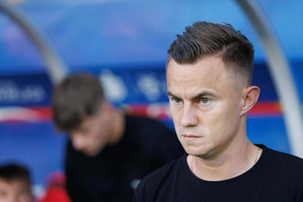 Mężczyzna z krótkimi, starannie zaczesanymi włosami i skupionym wyrazem twarzy, stojący na tle rozmytego stadionu lub ławki rezerwowych; w tle zamazane postacie.