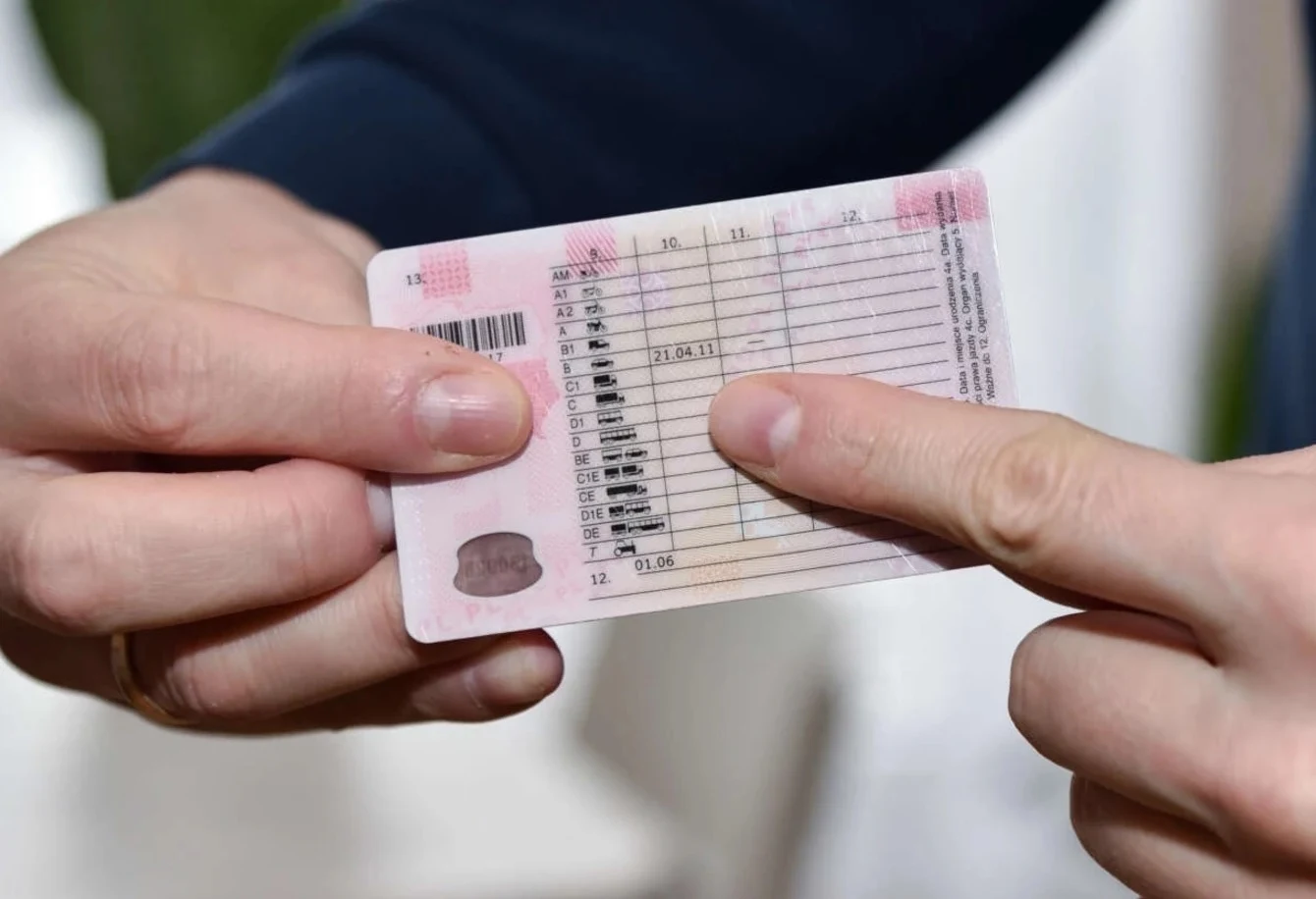 Według nowych przepisów dożywotnia utrata prawa jazdy będzie orzekana wobec kierowców, którzy złamali sądowy zakaz prowadzenia pojazdów Dwie dłonie trzymają dokument formatu karty, prawdopodobnie prawo jazdy, a jeden palec wskazuje na widoczne na nim miejsce z danymi.