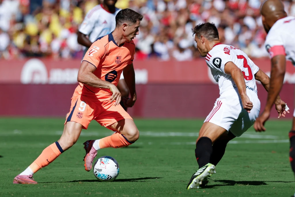 Robert Lewandowski w pojedynku z Cesarem Azpilicuetą Dwóch piłkarzy rywalizuje o piłkę podczas meczu piłki nożnej na zielonym boisku, jeden ubrany w pomarańczowy, a drugi w biały strój sportowy, w tle widoczna rozmyta publiczność.