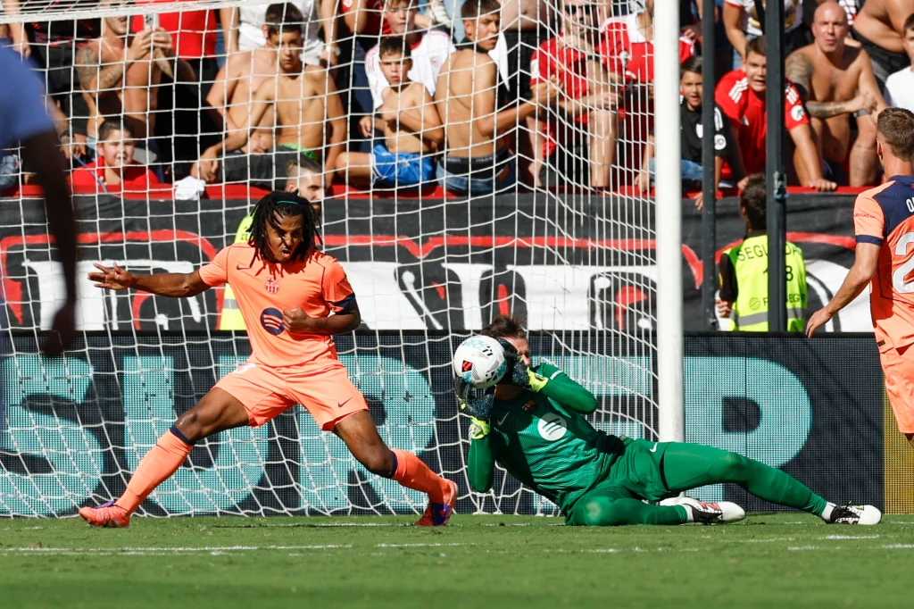 Piłkarz ubrany w pomarańczowy strój próbuje zdobyć bramkę, podczas gdy bramkarz w zielonym stroju interweniuje leżąc na murawie i chroniąc piłkę; w tle widoczny tłum kibiców.