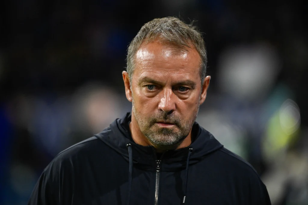 Hansi Flick Mężczyzna o poważnym wyrazie twarzy w sportowej bluzie na ciemnym, rozmytym tle