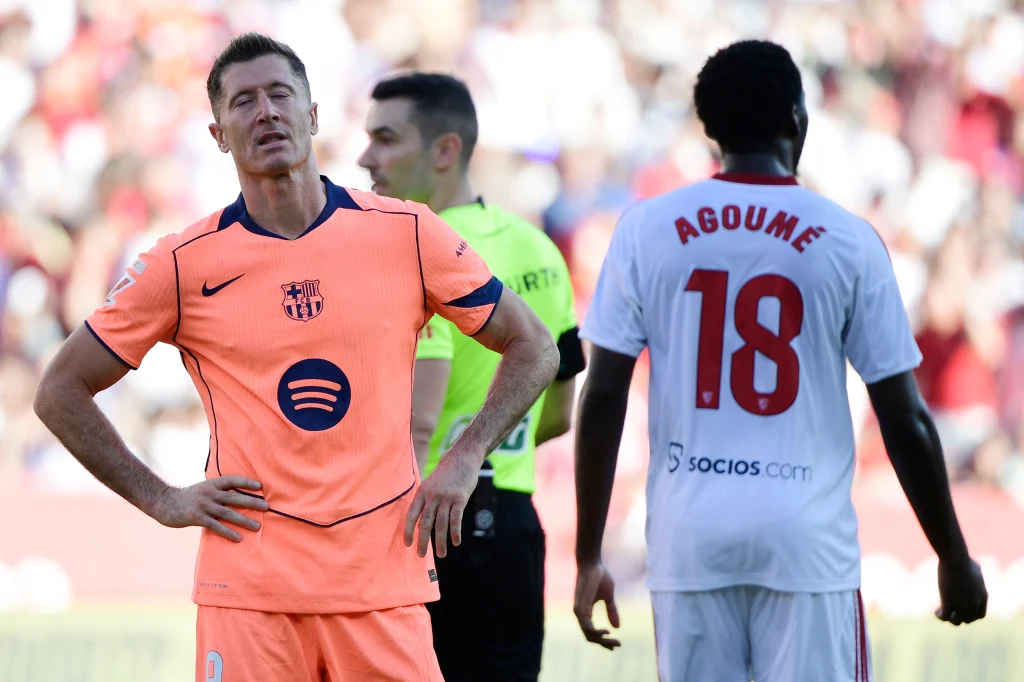 Robert Lewandowski Zawodnik w pomarańczowym stroju z logo FC Barcelony stoi z zamkniętymi oczami i rękami na biodrach, obok znajduje się zawodnik drużyny przeciwnej z numerem 18 na koszulce oraz sędzia w tle.