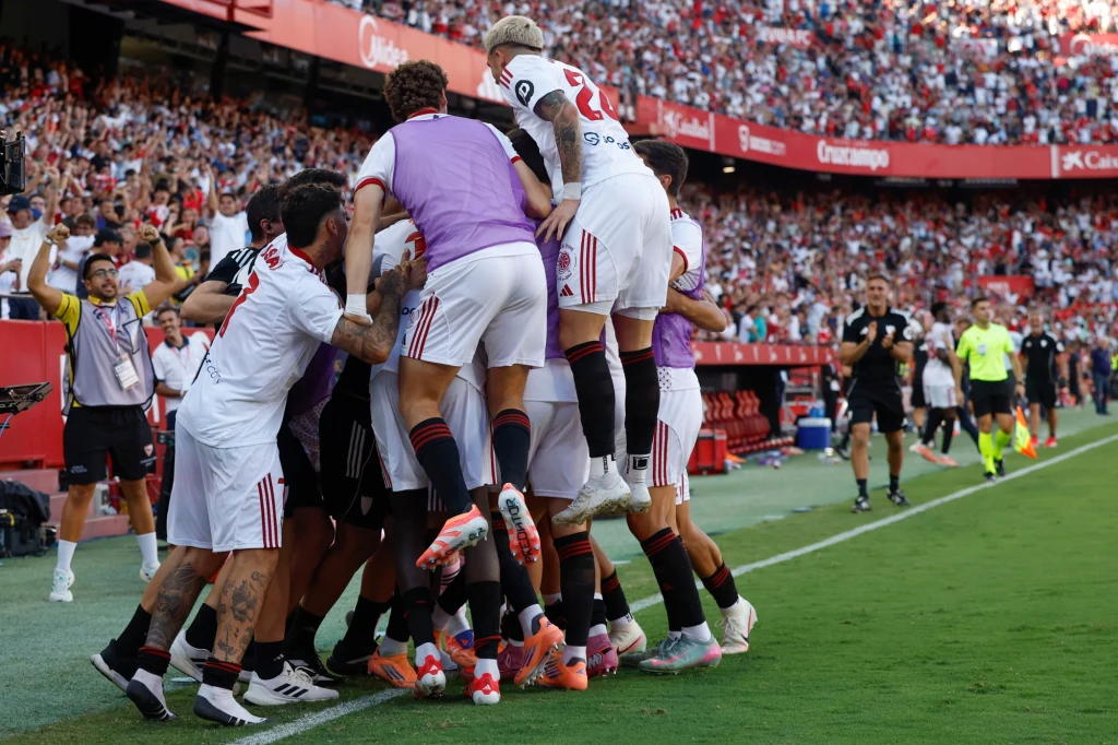 Ekipa z Sewilli wzięła srogi rewanż za dwa upokorzenia z poprzedniego sezonu Grupa piłkarzy świętuje razem przy linii bocznej boiska, tworząc stos z zawodników w białych i czarnych strojach, wokół pełne trybuny kibiców oraz sędziowie i członkowie sztabu w tle.