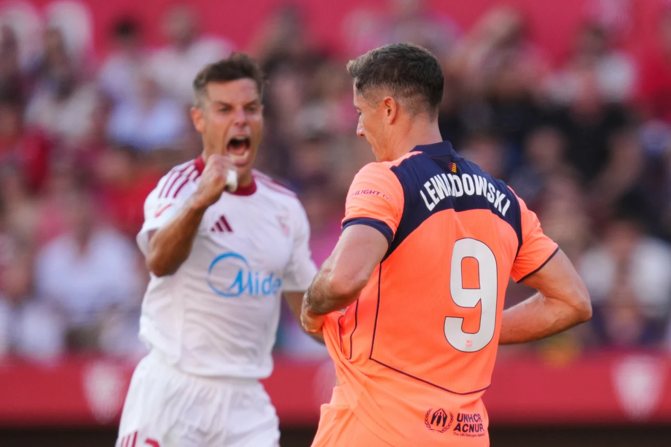 Dwóch piłkarzy na boisku podczas intensywnego momentu gry, jeden w białym stroju z emocjonalnym wyrazem twarzy kieruje gest w stronę drugiego zawodnika w pomarańczowej koszulce z numerem 9.