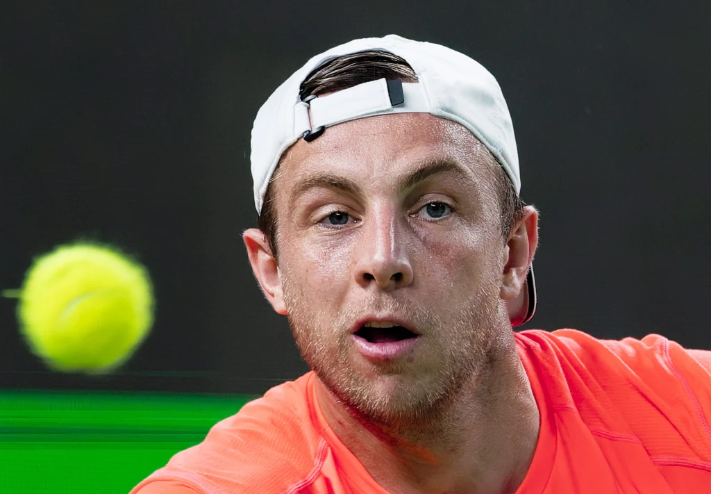 Zawodnik tenisa w intensywnym skupieniu podczas gry, ujęcie bliskie twarzy, z żółtą piłką tenisową widoczną na pierwszym planie. Sportowiec ubrany w jasną czapkę z daszkiem założoną tył na przód oraz pomarańczową koszulkę.