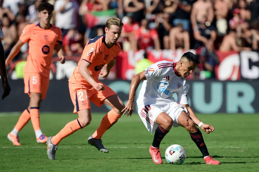 Dwóch piłkarzy walczy o piłkę na boisku podczas meczu, jeden z zawodników w białym stroju schyla się nagle jakby tracił równowagę, piłka znajduje się blisko jego stopy, w tle widoczna publiczność kibicująca drużynom.