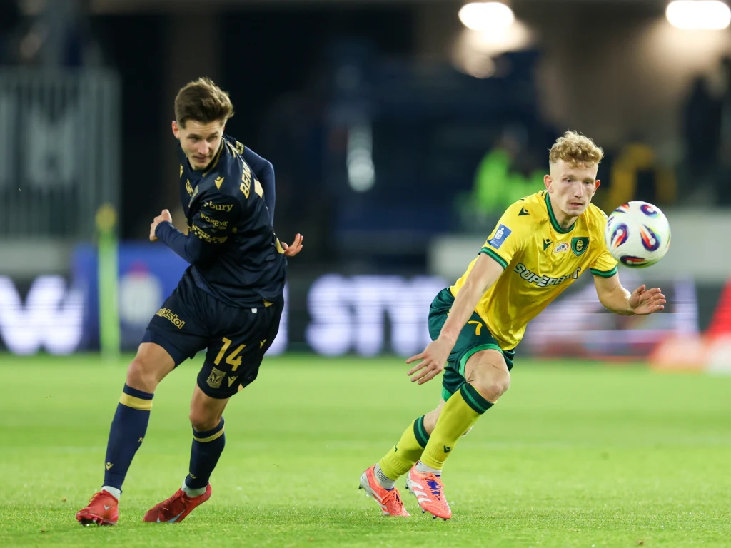 GKS Katowice - Lech Poznań Dwóch piłkarzy walczy o piłkę na boisku podczas meczu, jeden ubrany w granatowy, a drugi w żółty strój sportowy, w tle rozmazane trybuny stadionu