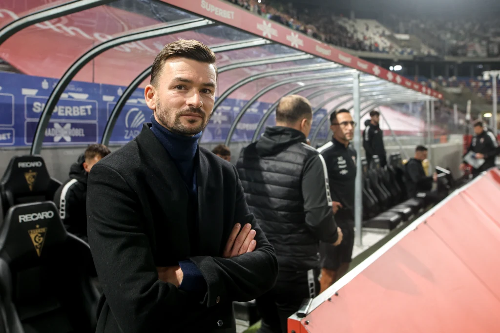 Michal Gasparik, trener aktualnego lidera Ekstraklasy Mężczyzna w ciemnym płaszczu stoi na stadionie przy ławce rezerwowych, w tle kilku członków sztabu trenerskiego oraz widownia stadionu piłkarskiego.
