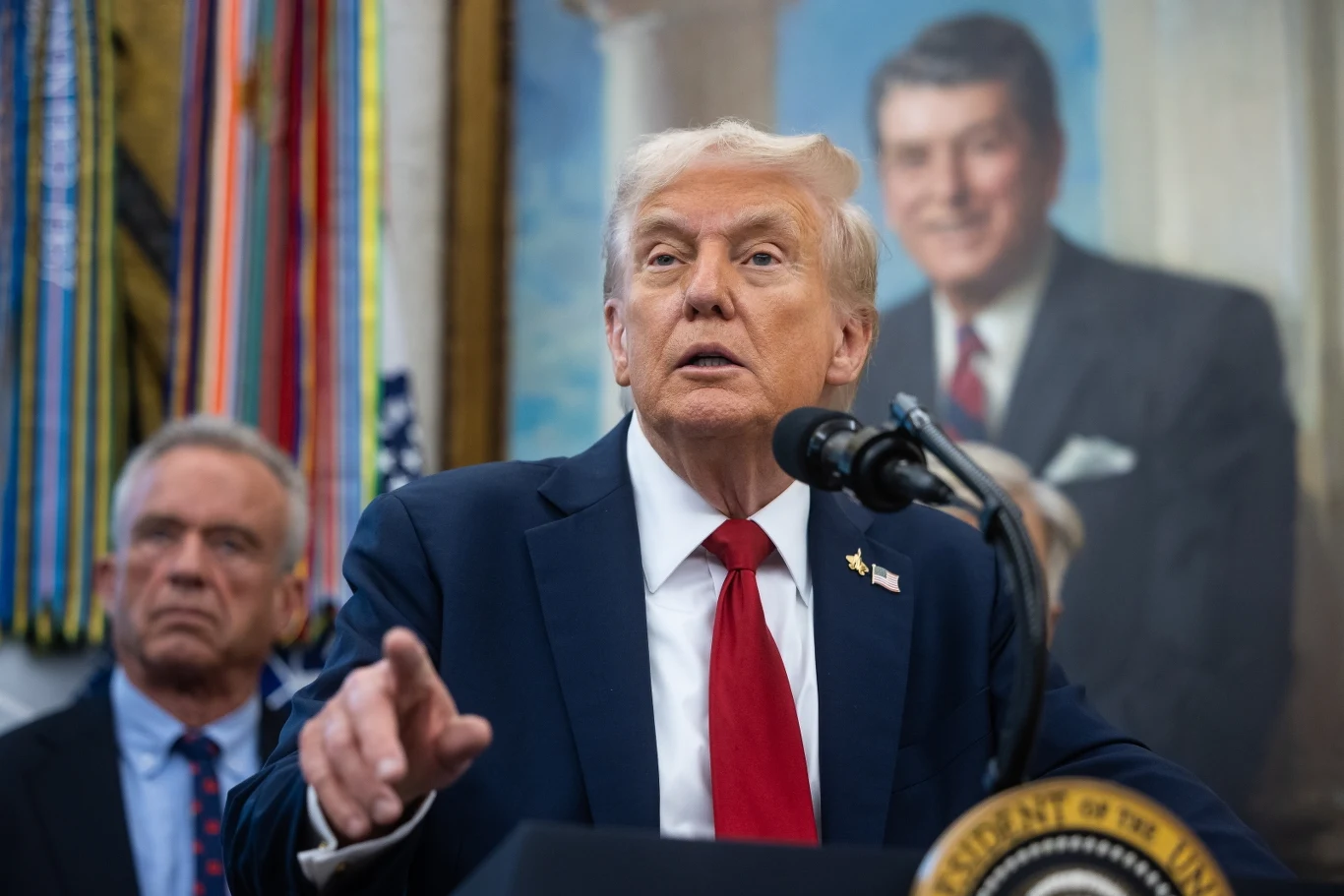 Prezydent USA Donald Trump Mężczyzna w garniturze z czerwonym krawatem przemawia przy mikrofonie w otoczeniu innych osób, w tle portret innego polityka oraz liczne sztandary wojskowe.