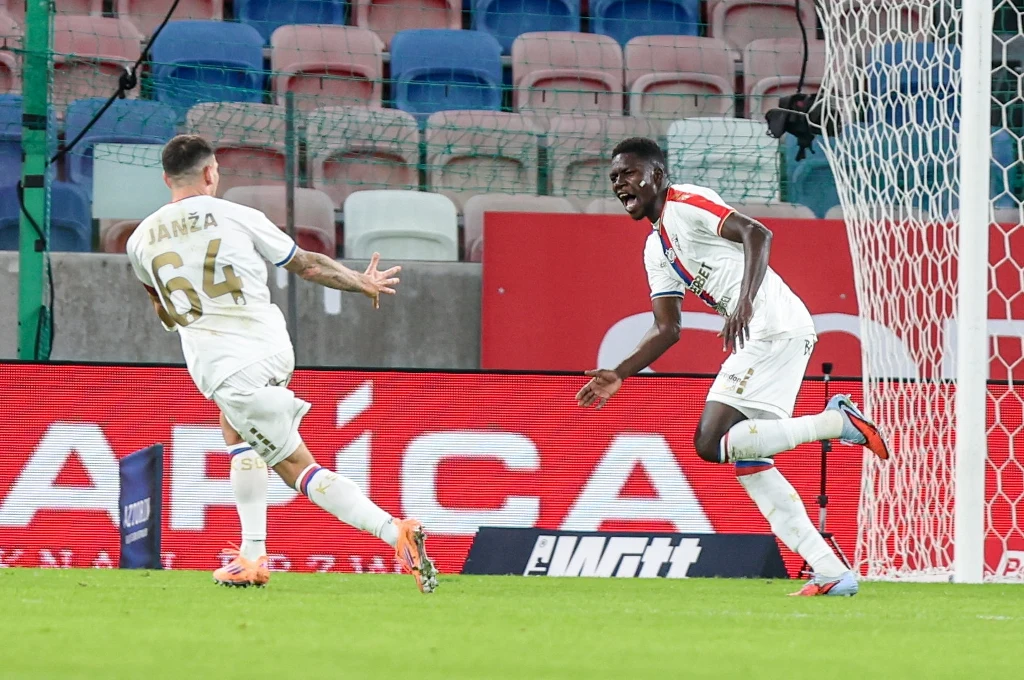 Bohaterem spotkania okazał się Ousmane Sow - dwie bramki i asysta na koncie Dwóch piłkarzy w białych strojach świętuje zdobycie bramki na stadionie, w tle puste trybuny oraz bramka z siatką.