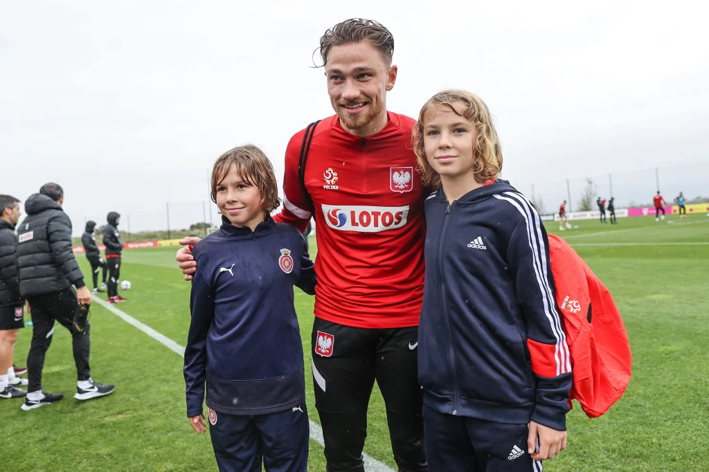 Zawodnik piłkarskiej reprezentacji Polski w czerwonym stroju sportowym pozuje na boisku z dwójką uśmiechniętych dzieci ubranych w sportowe bluzy, w tle widoczni są inni sportowcy oraz trawiasty stadion.