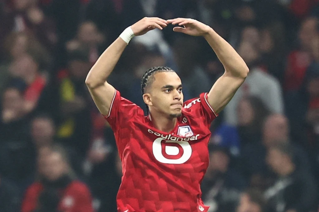 Piłkarz w czerwonym stroju z logo klubu wykonuje gest z rękami uniesionymi nad głową podczas meczu, w tle widownia stadionu