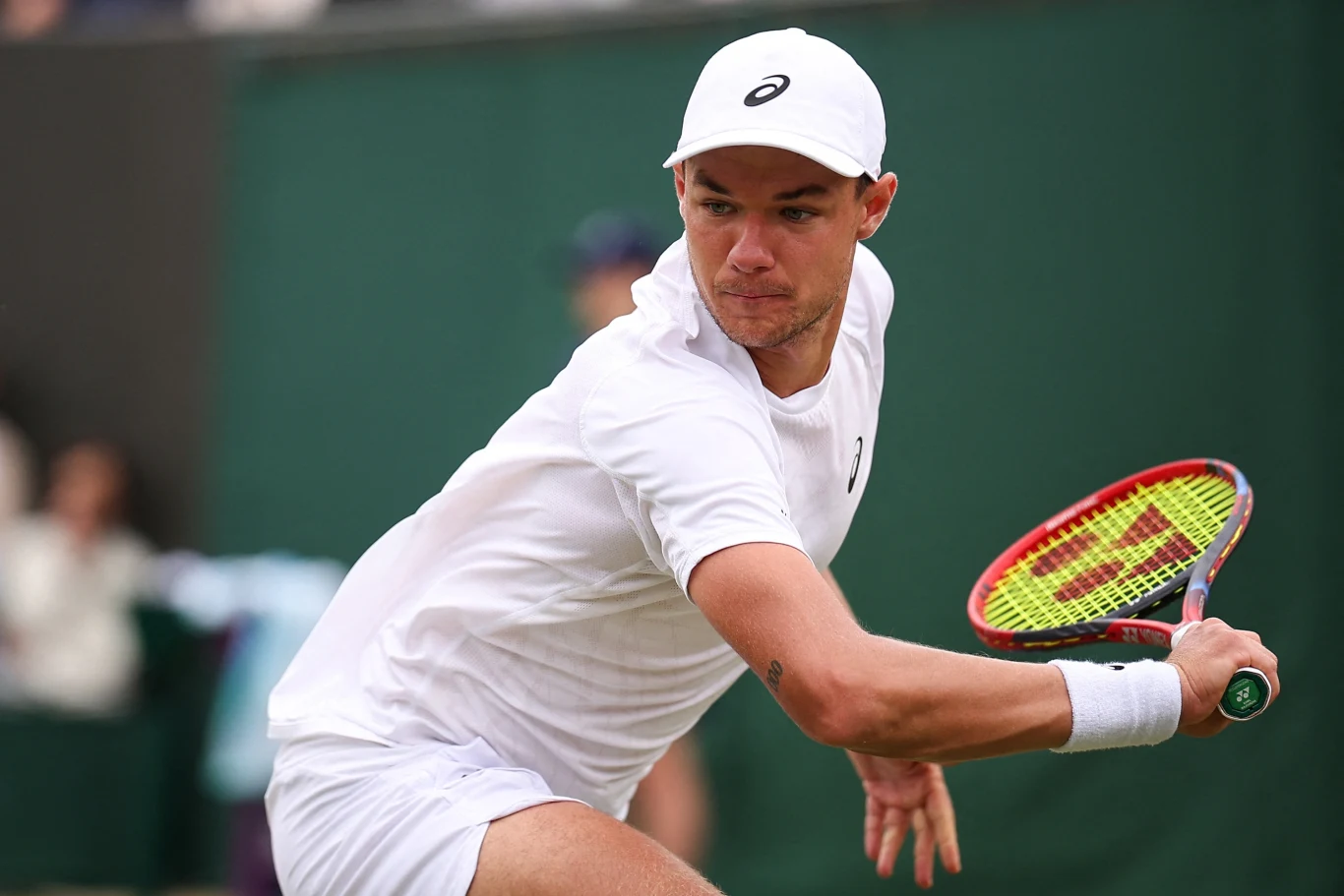 Tenisista w białym stroju skoncentrowany na grze podczas dynamicznego uderzenia rakietą tenisową, w tle rozmyte postacie kibiców.