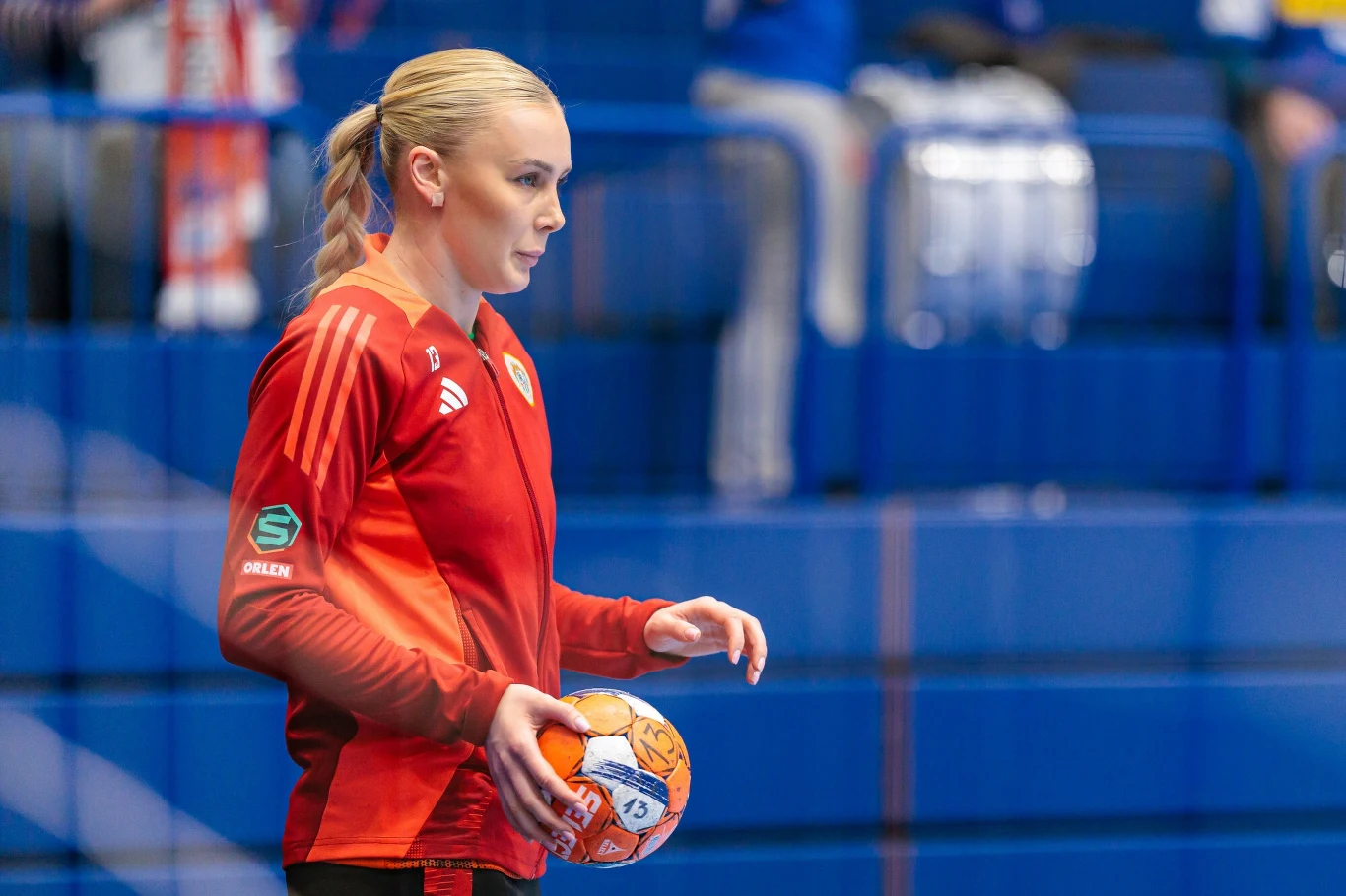 Kobieta w sportowym stroju w czerwonych barwach trzyma piłkę ręczną, znajduje się na hali sportowej z niebieskimi trybunami w tle