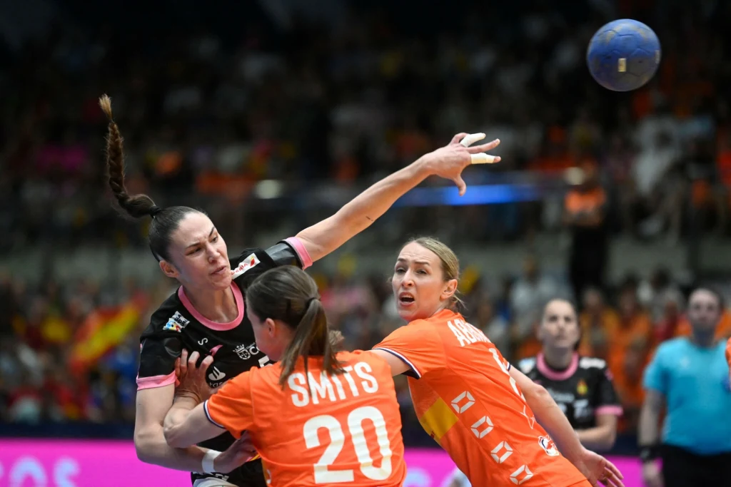 María Prieto O'Mullony (rzuca piłką) Zacięta walka między zawodniczkami podczas meczu piłki ręcznej, jedna zawodniczka w czarnym stroju próbuje zdobyć przewagę, podczas gdy dwie przeciwniczki w pomarańczowych strojach starają się ją powstrzymać, piłka unosząca się w powietrzu.