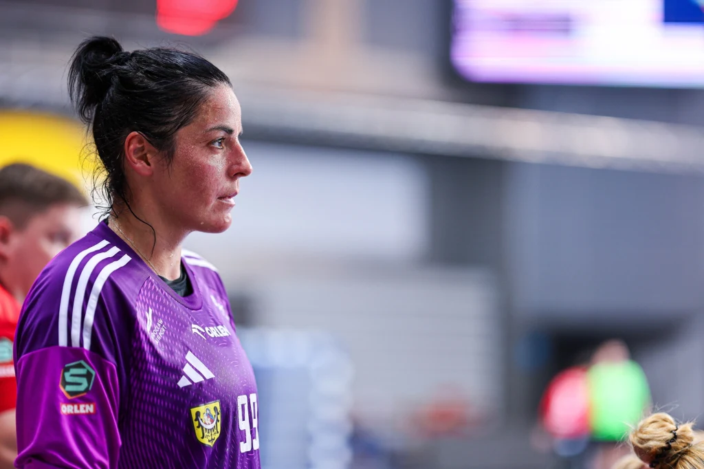 Zbliżenie na zawodniczkę drużyny piłki ręcznej w fioletowym sportowym stroju z numerem 99, skupioną podczas rozgrywek sportowych w hali.