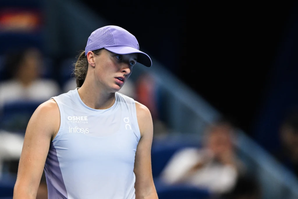 Kobieta w sportowym stroju i czapce z daszkiem znajduje się na korcie tenisowym, patrzy w bok z lekko zmęczonym wyrazem twarzy, w tle rozmyte postacie widzów.