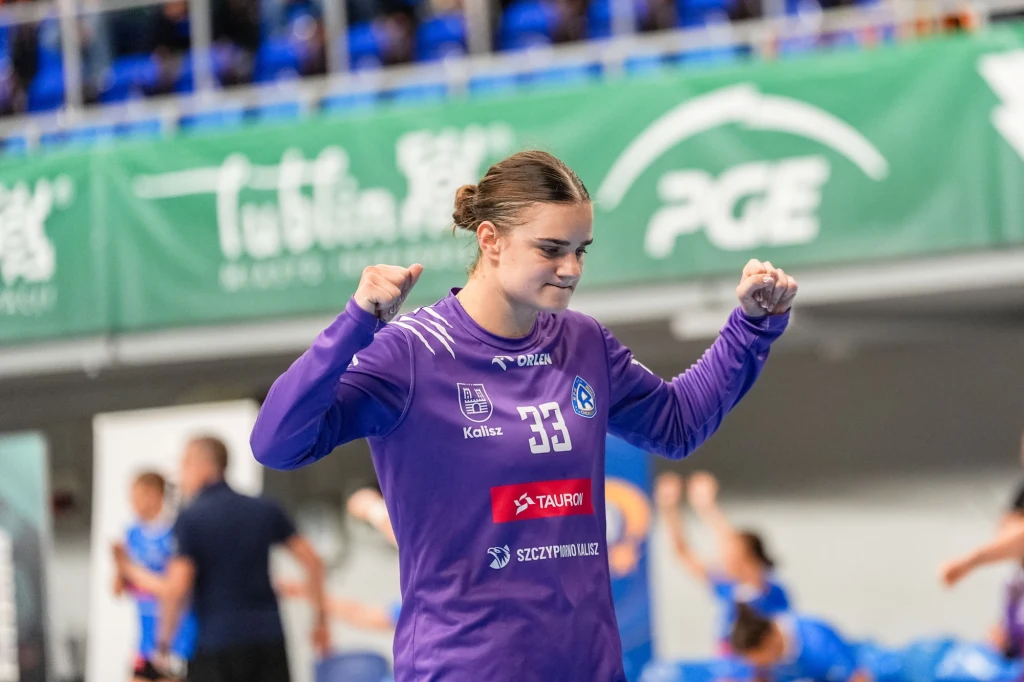 Zawodniczka drużyny piłki ręcznej ubrana w fioletowy strój z numerem 33 podczas sportowych emocji na hali, z uniesionymi rękami w geście radości, w tle widoczni kibice i fragmenty trybun.